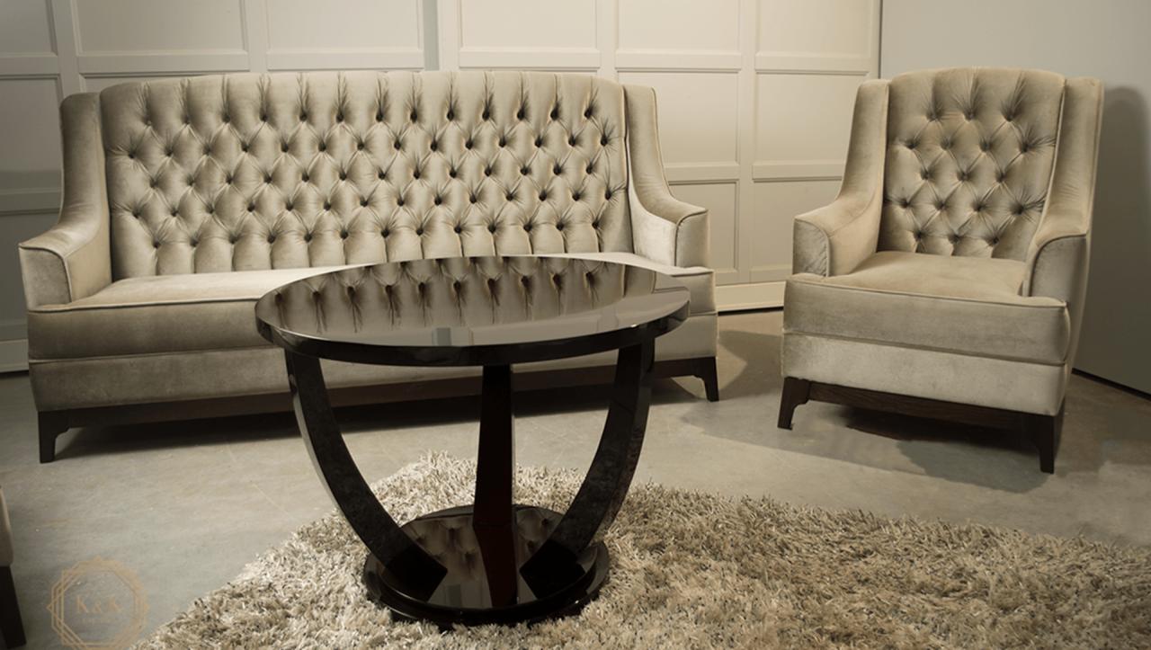 Beżowy, pikowany zestaw wypoczynkowy: sofa i fotel, z okrągłym, czarnym stolikiem kawowym na tle białej ściany z panelami i puszystego dywanu.