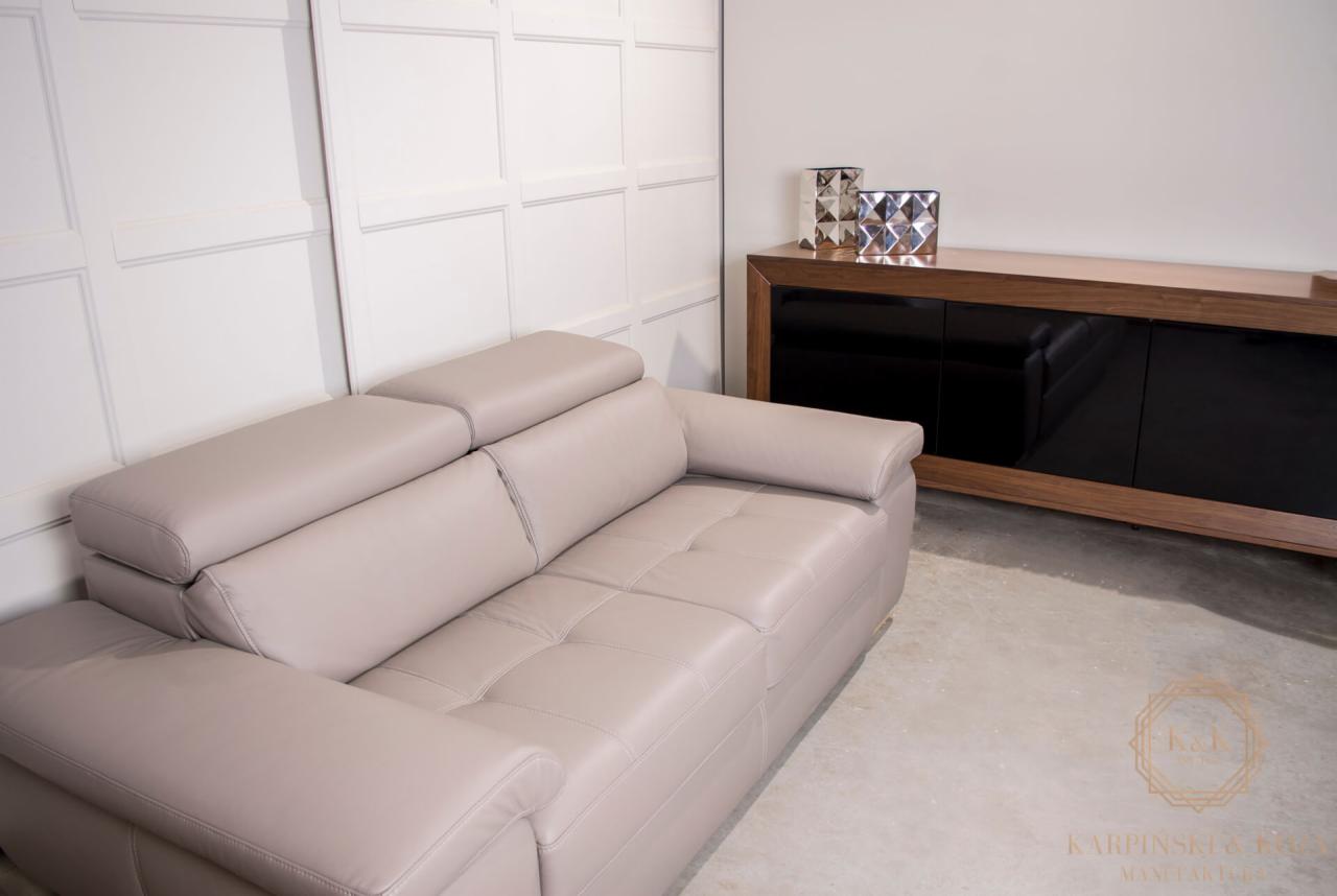 Jasnoszara, skórzana sofa z regulowanymi zagłówkami, ustawiona w nowoczesnym wnętrzu z białą, panelową ścianą i ciemnobrązową komodą z czarnymi frontami.