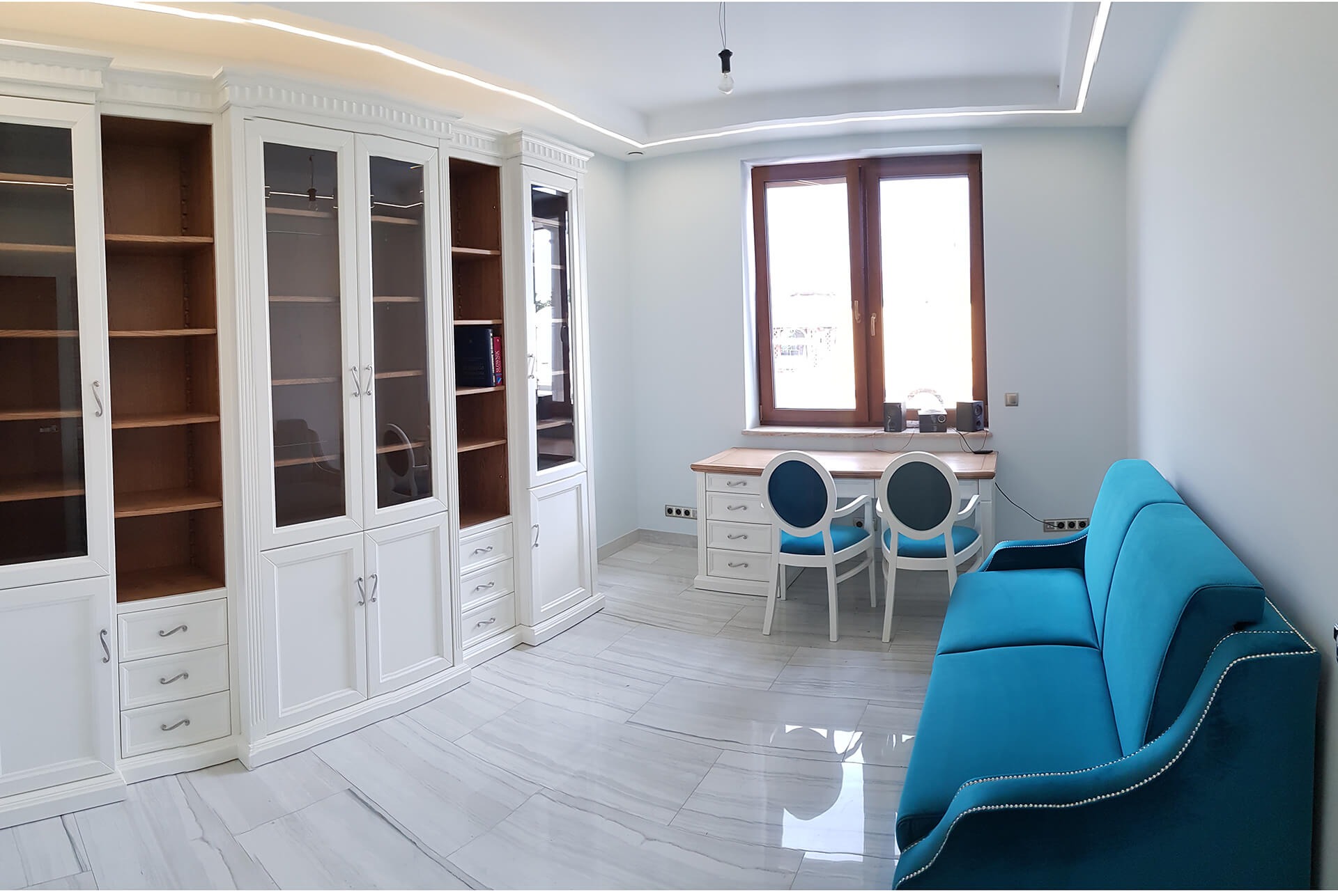 Jasne wnętrze gabinetu z białą zabudową biblioteczną, biurkiem z krzesłami i turkusową sofą na marmurowej podłodze.