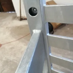 Fragment metalowej konstrukcji pomalowanej na szaro, widoczny spaw i otwór montażowy, w tle betonowa podłoga i częściowo widoczny karton.