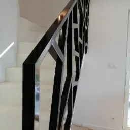 Nowoczesna balustrada schodowa z czarnych i srebrnych metalowych profili o geometrycznym wzorze, widok z dołu na białe schody i ścianę.