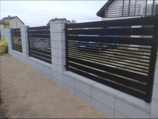 Nowoczesne ogrodzenie z poziomych, czarnych paneli metalowych osadzonych na szarym, betonowym murku z filarami, widok z zewnątrz posesji.