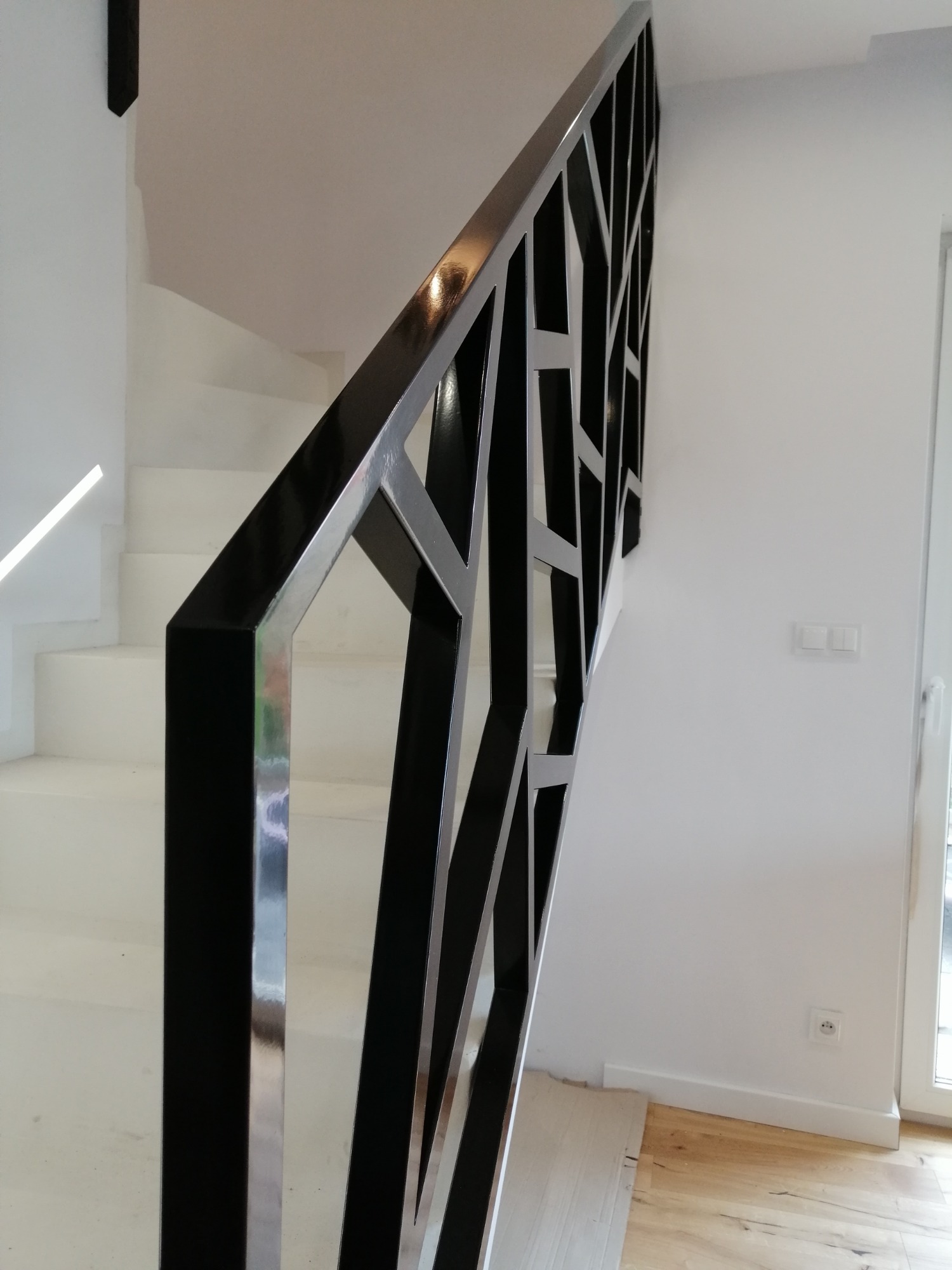 Nowoczesna balustrada schodowa z czarnych i srebrnych metalowych profili o geometrycznym wzorze, widok z dołu na białe schody i ścianę.