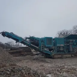 Turkusowa kruszarka gruzu na gąsienicach przesypuje urobek na hałdę, w tle pomarańczowa koparka na terenie budowy.