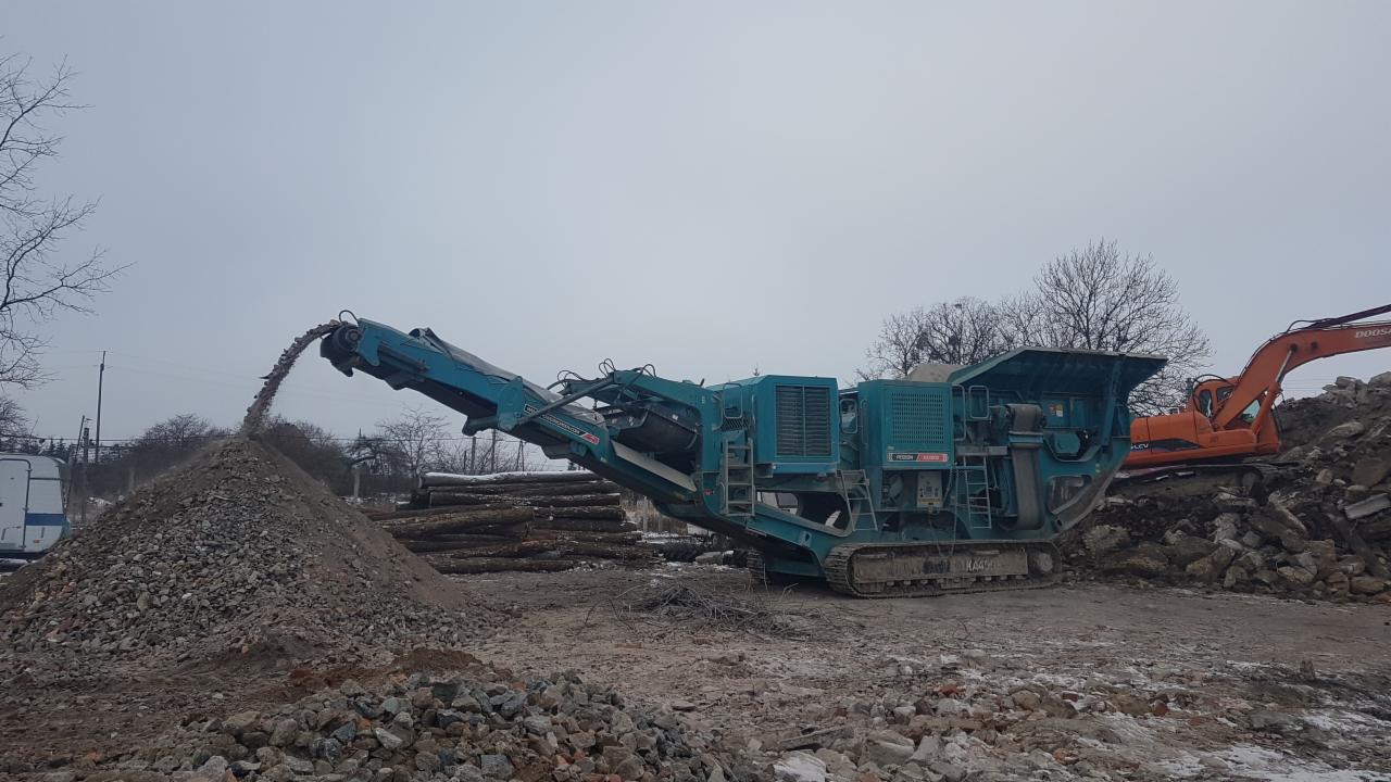 Turkusowa kruszarka gruzu na gąsienicach przesypuje urobek na hałdę, w tle pomarańczowa koparka na terenie budowy.