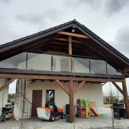 MARIUSZ SŁOMIŃSKI - Dwukondygnacyjny budynek z drewnianą konstrukcją i szklaną balustradą na górnym balkonie, widok z poziomu podwórka, z widocznymi narzędziami ogrodniczymi i zabawkami dziecięcymi w tle.