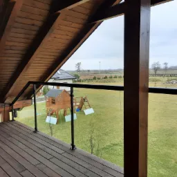 Zadaszony taras z drewnianą podłogą i szklaną balustradą, widok na zielony ogród z domkiem dla dzieci i placem zabaw w tle.