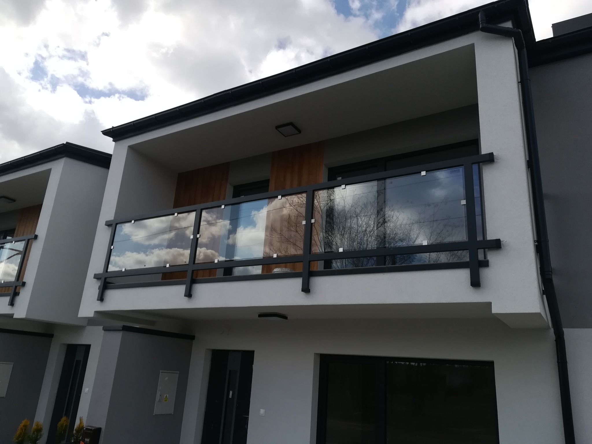 Nowoczesna balustrada szklana z czarnymi elementami konstrukcyjnymi na balkonie bliźniaka, odbijająca chmury i drzewa.