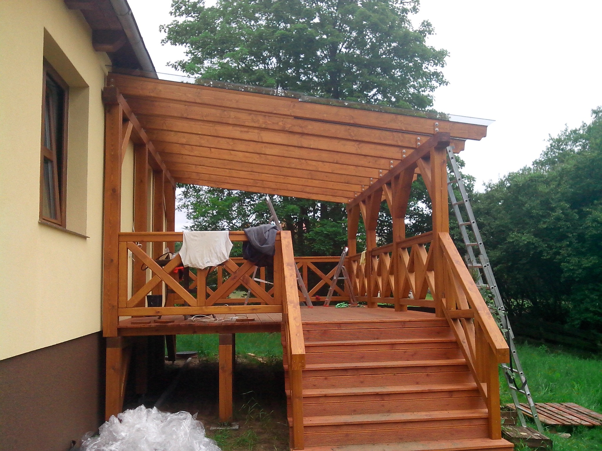Drewniana zabudowa balkonu z zadaszeniem, widoczne schody, balustrada z krzyżowym wzorem, drabina oparta o konstrukcję, częściowo wykończona, z elementami budowlanymi w pobliżu.