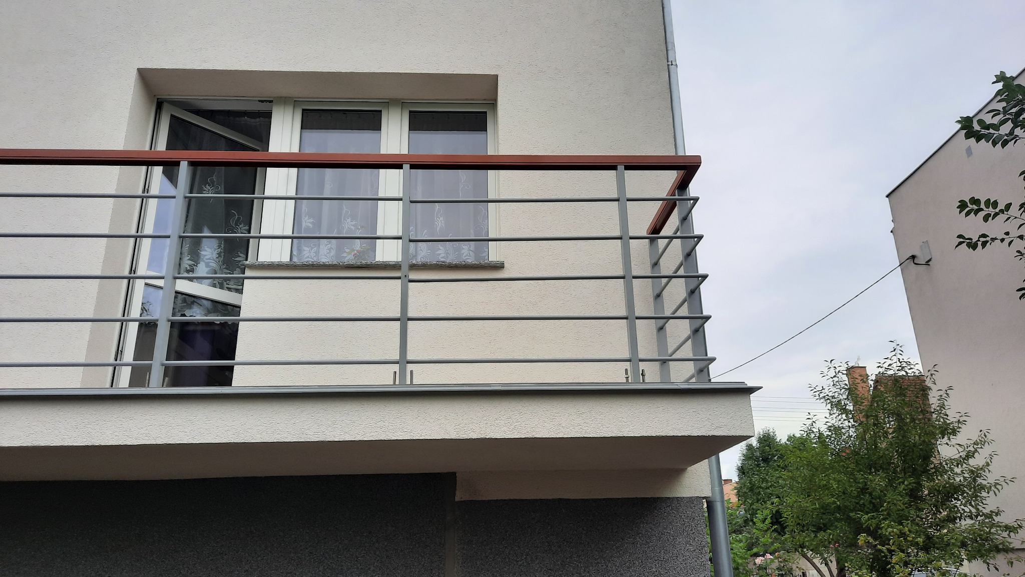 Balkon z szarą metalową balustradą z drewnianym poręczą na tle jasnej elewacji budynku z oknami.