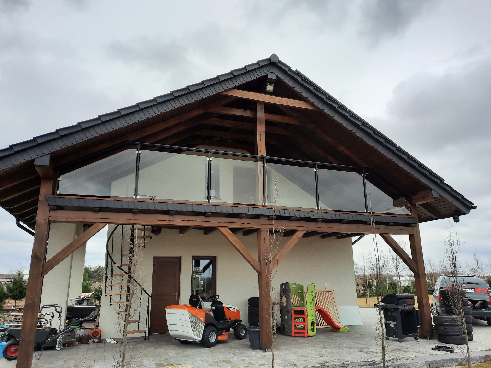 Dwukondygnacyjny budynek z drewnianą konstrukcją i szklaną balustradą na górnym balkonie, widok z poziomu podwórka, z widocznymi narzędziami ogrodniczymi i zabawkami dziecięcymi w tle.