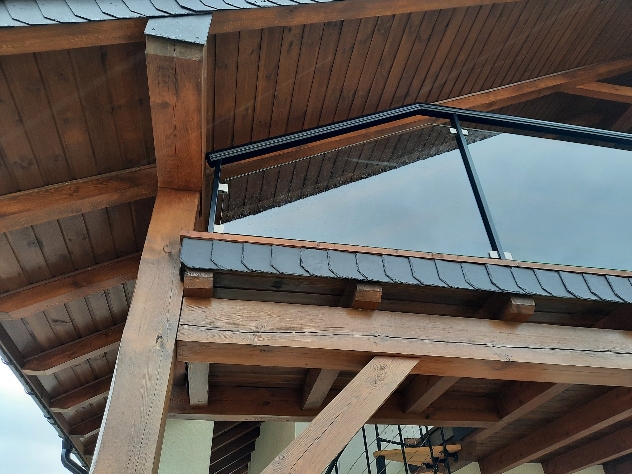 Szklana balustrada na drewnianej konstrukcji balkonu z widocznym fragmentem dachu pokrytego łupkiem.