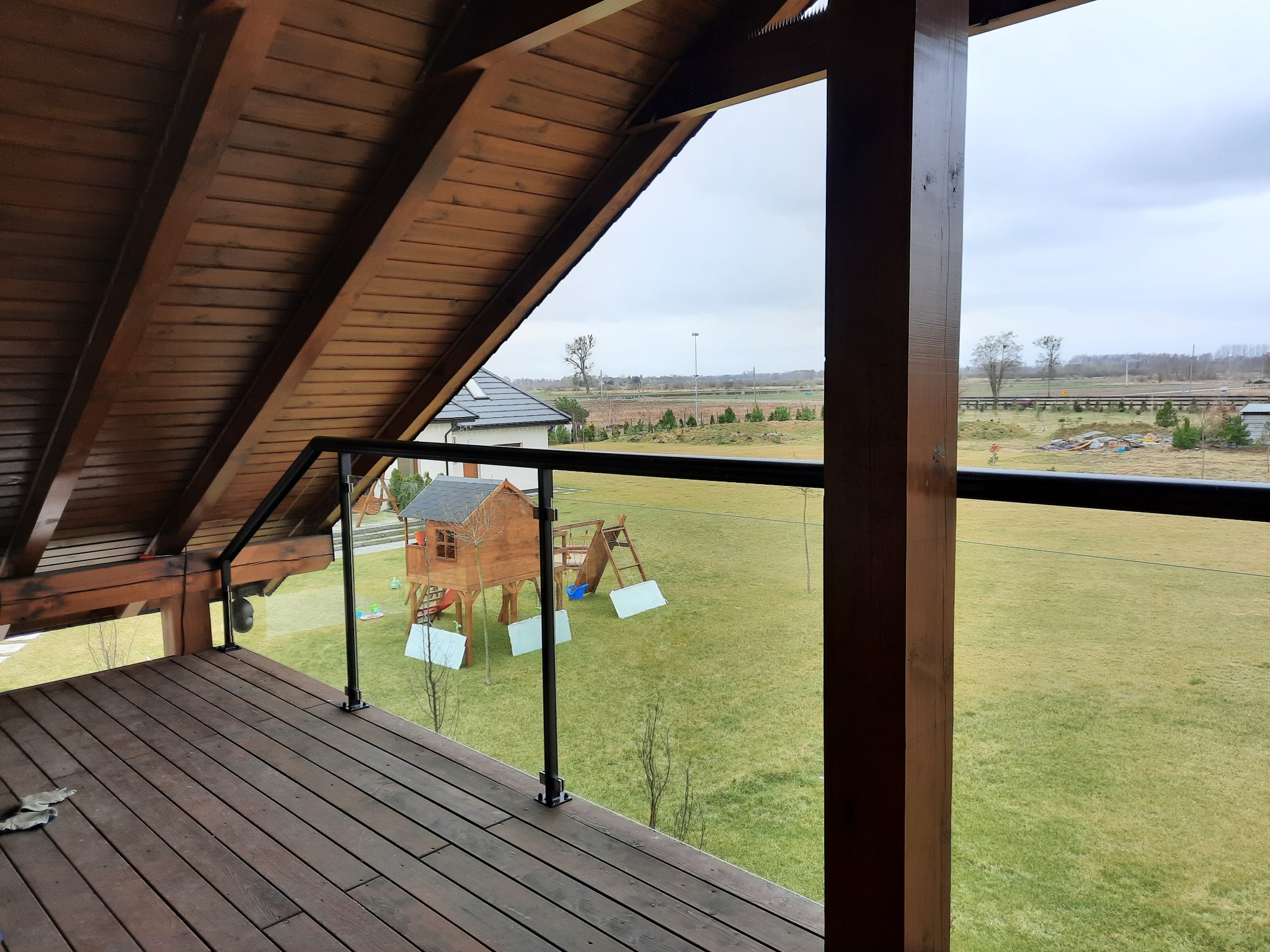 Zadaszony taras z drewnianą podłogą i szklaną balustradą, widok na zielony ogród z domkiem dla dzieci i placem zabaw w tle.