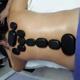 Kobieta leżąca na brzuchu z ułożonymi na plecach gorącymi, czarnymi kamieniami podczas zabiegu relaksacyjnego w gabinecie.