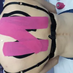 Plecy kobiety z aplikacją różowych taśm kinesiotaping, widoczny fragment biustonosza i dżinsów, obok leży rolka taśmy.
