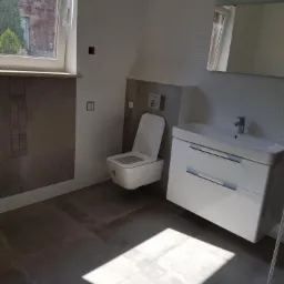 Nowoczesna łazienka z podwieszaną toaletą, białą szafką z umywalką i szarą glazurą, oświetlona naturalnym światłem z okna.