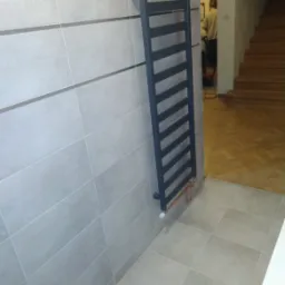 Zamontowany, grafitowy grzejnik drabinkowy na szarej, poziomej ścianie z płytek w łazience. Widoczne miedziane rurki doprowadzające wodę. W tle fragment schod&oacute;w i podłogi z parkietu.