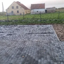 Ułożona kostka brukowa w szarym odcieniu, przylegająca do powierzchni wysypanej drobnym, szarym żwirem, z widoczną siatką wzmacniającą podłoże, na tle wiejskiego krajobrazu z domami i ogrodzeniem.
