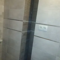 Wykończona łazienka z szarymi płytkami o prostokątnym kształcie, dekoracyjną wstawką imitującą drewno i podwójnym gniazdkiem elektrycznym.