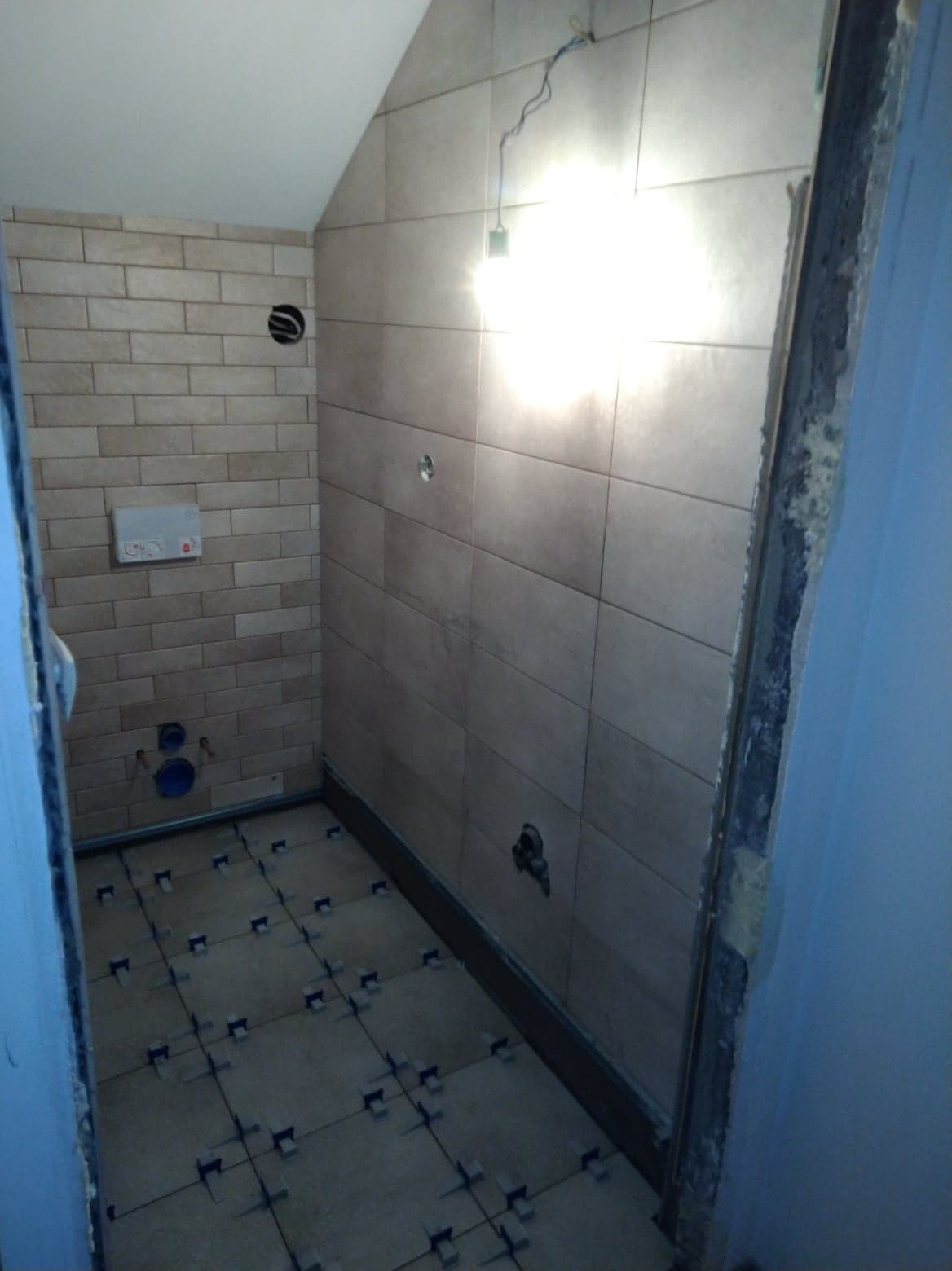 Łazienka w trakcie remontu, widoczne płytki na ścianach i podłodze z krzyżykami dystansowymi, instalacja sanitarna w ścianie, surowa krawędź ściany przy drzwiach.