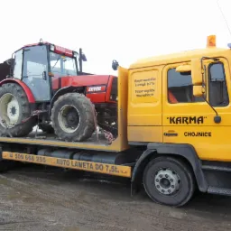Żółta laweta z napisem 'KARMA Chojnice' przewozi czerwony ciągnik Zetor. Laweta oferuje wynajem podnośników koszowych do 27m oraz transport ciągników i maszyn rolniczych.
