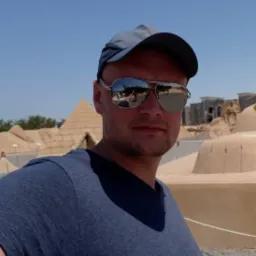 Mężczyzna w okularach przeciwsłonecznych i czapce z daszkiem na tle stylizowanej scenerii egipskiej z piramidami i sfinksami.