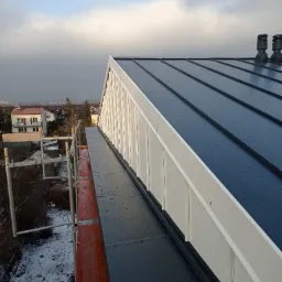 Nowy, ciemny dach z blachy trapezowej z dwoma kominkami wentylacyjnymi, widoczny z rusztowania na tle zabudowy miejskiej i pochmurnego nieba.