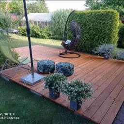 Drewniany taras z meblami ogrodowymi: wiszące krzesło, pufy w liście i kwiaty w donicach, otoczony zielenią trawnika i żywopłotu.