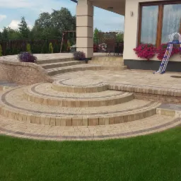 Okrągłe schody i taras z beżowej kostki brukowej z ciemniejszymi obwódkami, prowadzące do domu z kwiatami w oknach i rozstawioną drabiną.