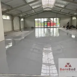 Świeżo wylana, szara posadzka żywiczna w hali przemysłowej z widocznymi refleksami światła i ekipą montażową w tle, logo firmy w prawym dolnym rogu.