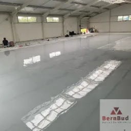 Aplikacja szarej, samopoziomującej wylewki w hali przemysłowej, widoczny pracownik z maszyną szlifującą, odbicie okien w mokrej powierzchni, logo firmy w prawym dolnym rogu.