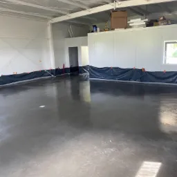 Świeżo wylana, błyszcząca posadzka w hali przemysłowej, zabezpieczona czarną folią z pomarańczowymi klipsami wzdłuż ścian.