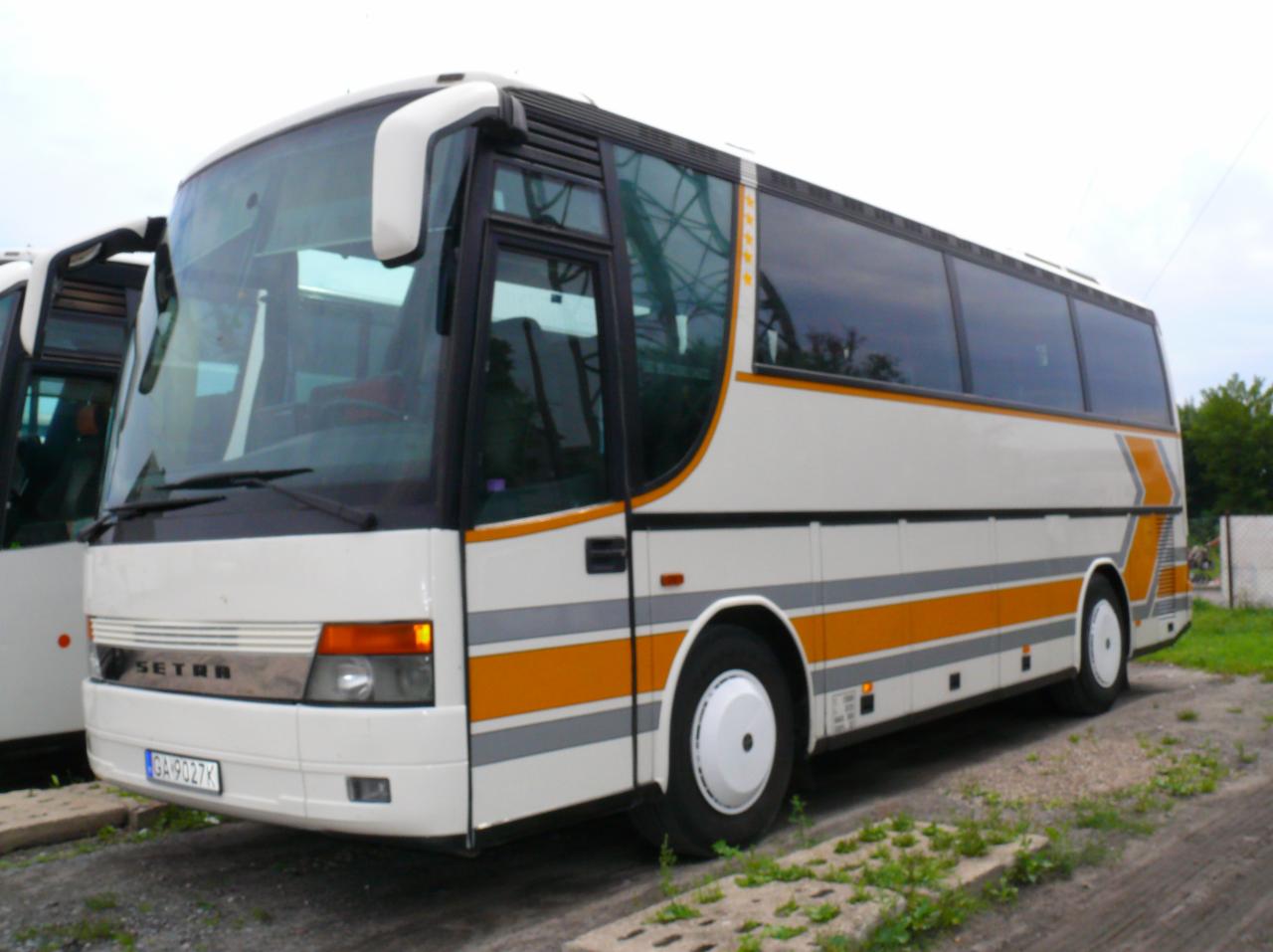 Biało-pomarańczowy autokar Setra z widoczną tablicą rejestracyjną, zaparkowany na betonowych płytach z zielenią.