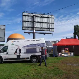 Reklamowy van z grafiką przedstawiającą okna, bramy i rolety, zaparkowany na trawniku obok stoiska z popcornem i watą cukrową, w tle billboard i budynki miejskie.