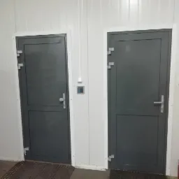 Dwie szare, stalowe bramy przemysłowe z białymi ościeżnicami w jasnym pomieszczeniu z panelami ściennymi, widoczna szufla do śniegu i wycieraczka.