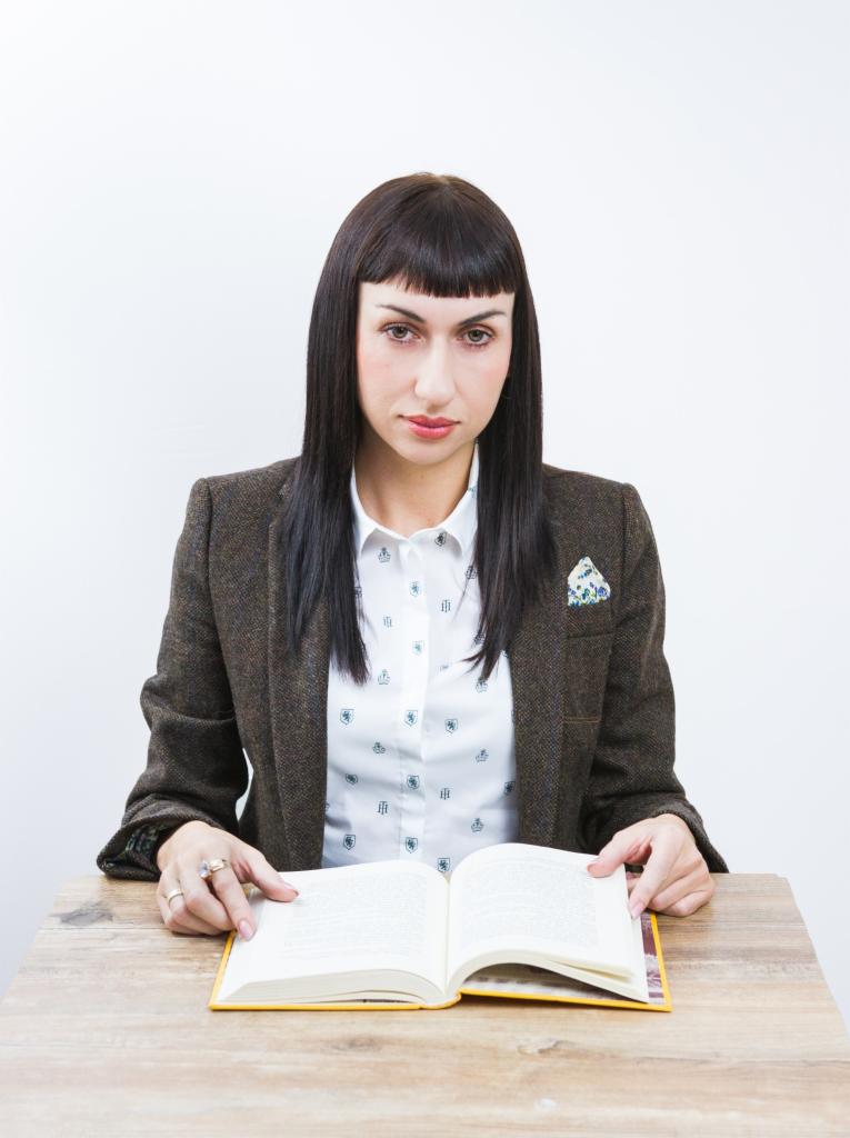 Kobieta w tweedowej marynarce i wzorzystej koszuli, siedząca za biurkiem i czytająca otwartą książkę, z poważnym wyrazem twarzy.