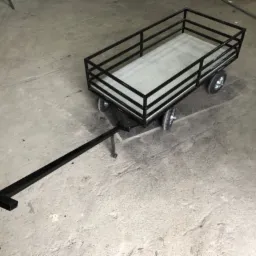 Czarny, metalowy wózek transportowy na czterech kołach z długim dyszlem, stojący na betonowej posadzce.