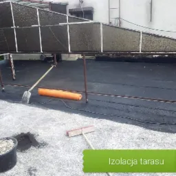 Prace izolacyjne na tarasie: widok na pokryty czarnym materiałem dach, z widoczną łopatą, rolką izolacji i wiadrem z zaprawą, w tle konstrukcja z metalowych profili.