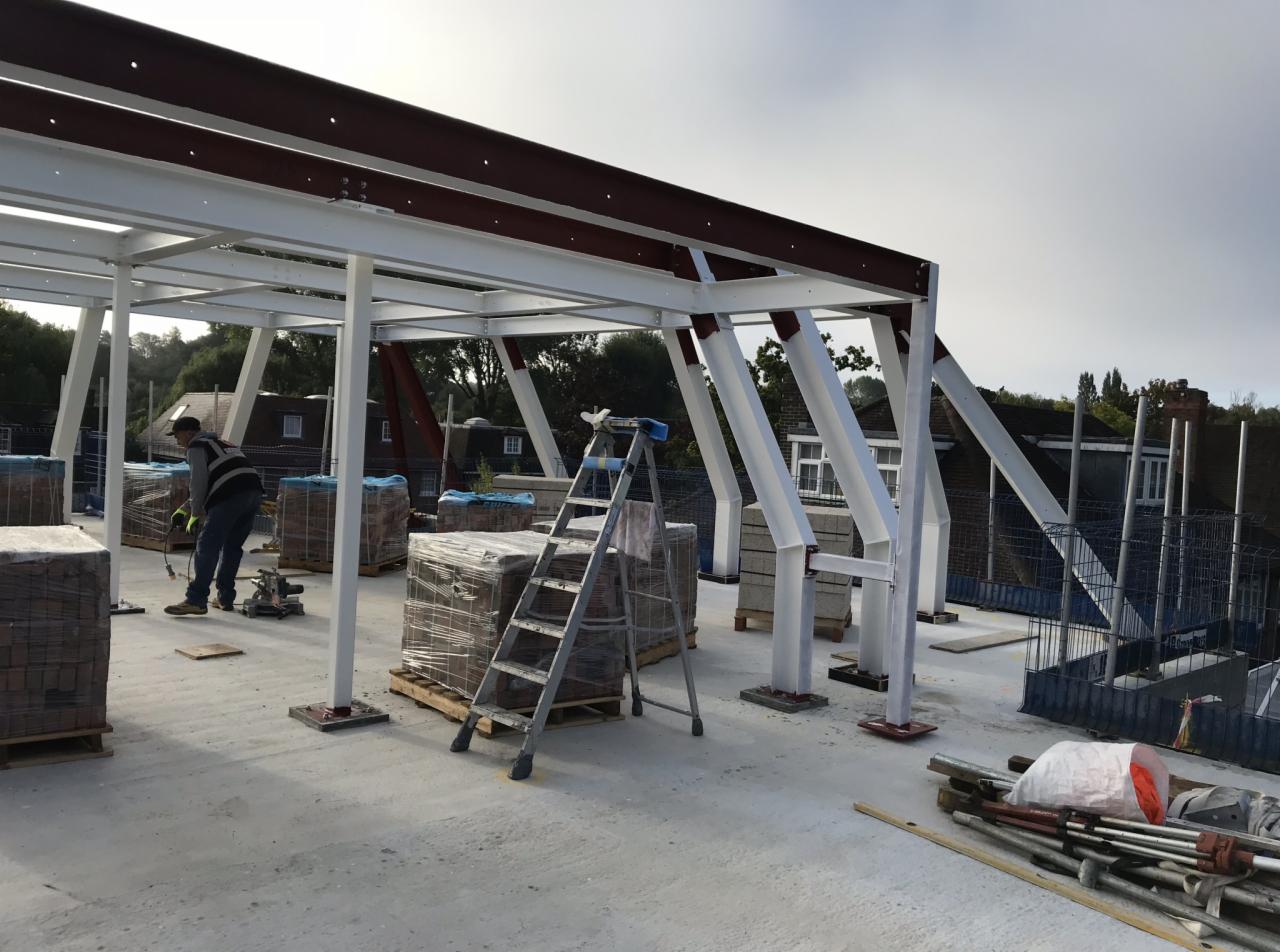 Konstrukcja stalowa dachu w trakcie budowy w Londynie, widoczne białe słupy, brązowe belki, palety z materiałami budowlanymi i drabina.