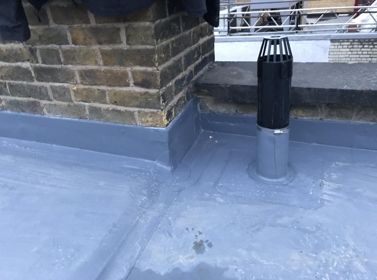 Szczelne połączenie muru kominowego z płaskim dachem pokrytym szarą izolacją, widoczna rura wentylacyjna z czarnym kominkiem wentylacyjnym.