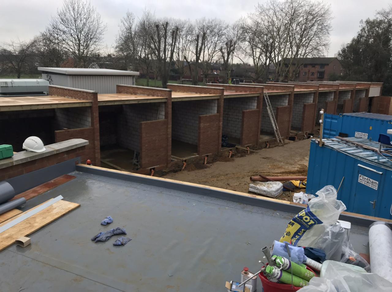 Budowa szeregu garaży z cegły i bloczków, widok z góry na płaski dach, częściowo zadaszone konstrukcje z widocznymi elementami drewnianymi, teren budowy z kontenerami i narzędziami w tle.