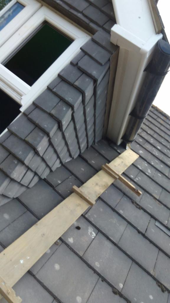 Szare dachówki ułożone wokół okna dachowego i rynny, zabezpieczone drewnianą deską podczas montażu.