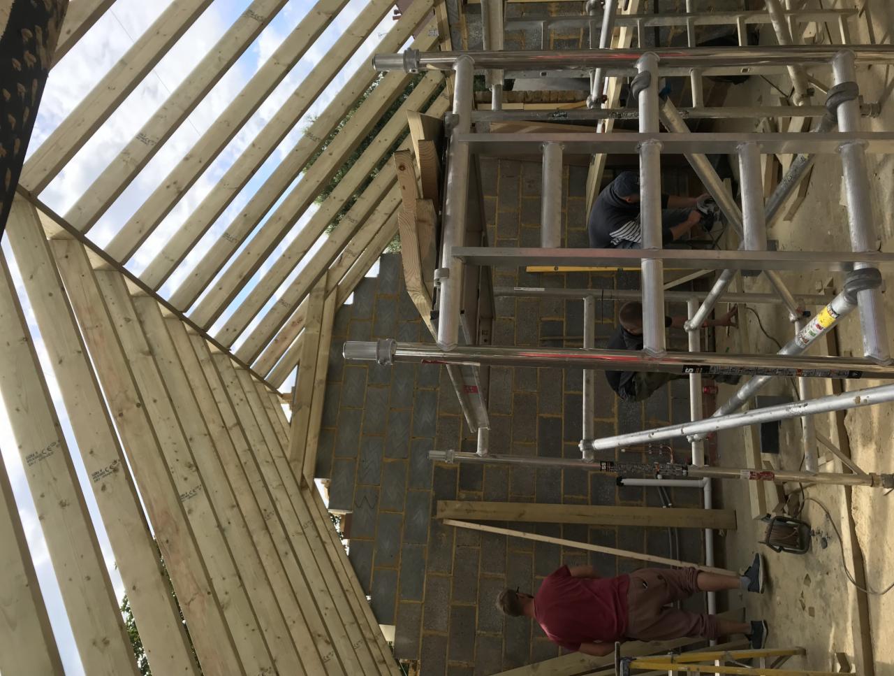 Montaż konstrukcji drewnianej dachu z użyciem rusztowania, widoczni pracownicy w trakcie pracy, surowy stan, cegły silikatowe.