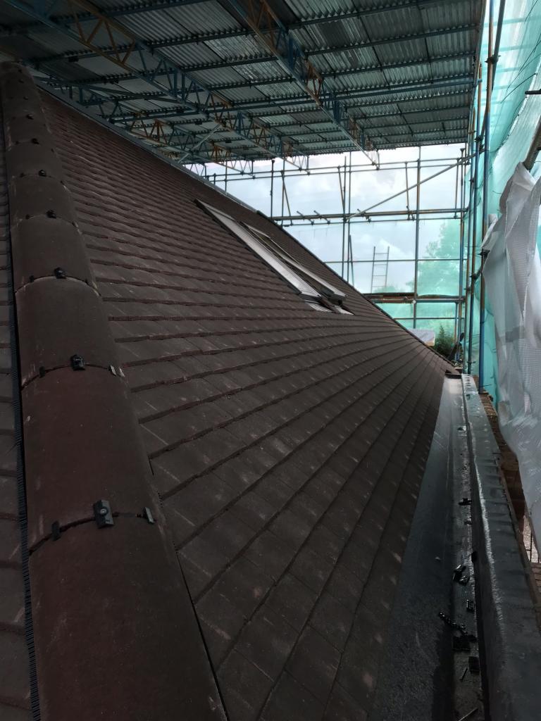 Ciemna dachówka ceramiczna na stromym dachu z oknem połaciowym, widoczne rusztowanie zabezpieczające prace dekarskie.