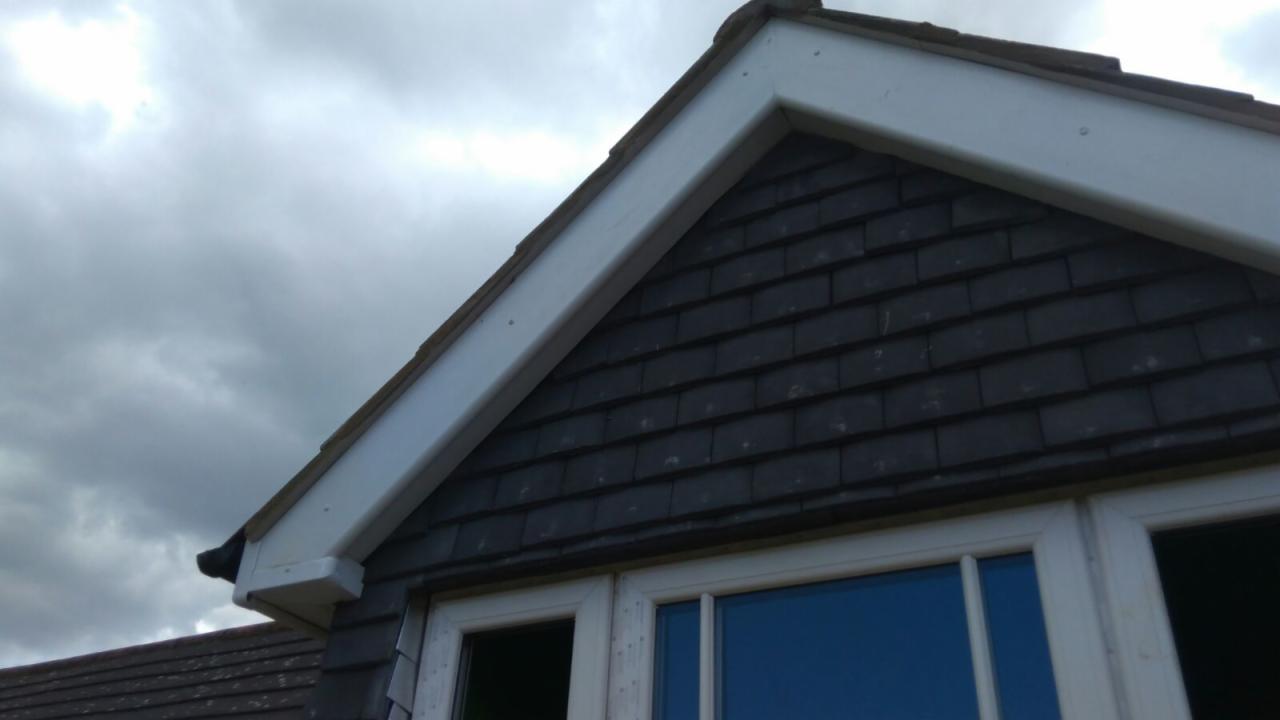 Fragment dachu z szarą dachówką łuskową, białą podbitką i oknem w białej ramie na tle pochmurnego nieba.