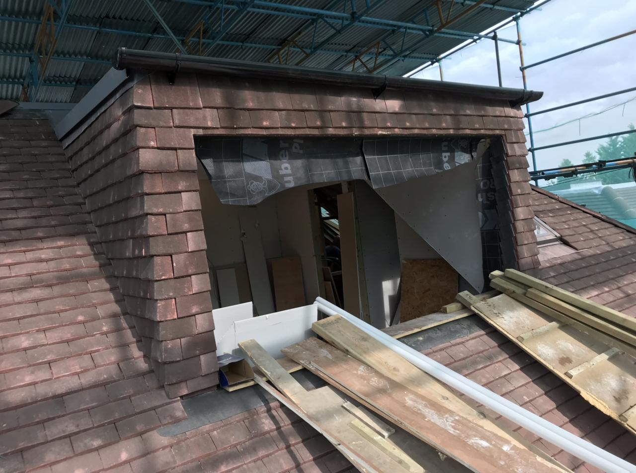 Fragment dachu z oknem połaciowym w trakcie montażu, widoczne deski, membrana i rusztowanie.