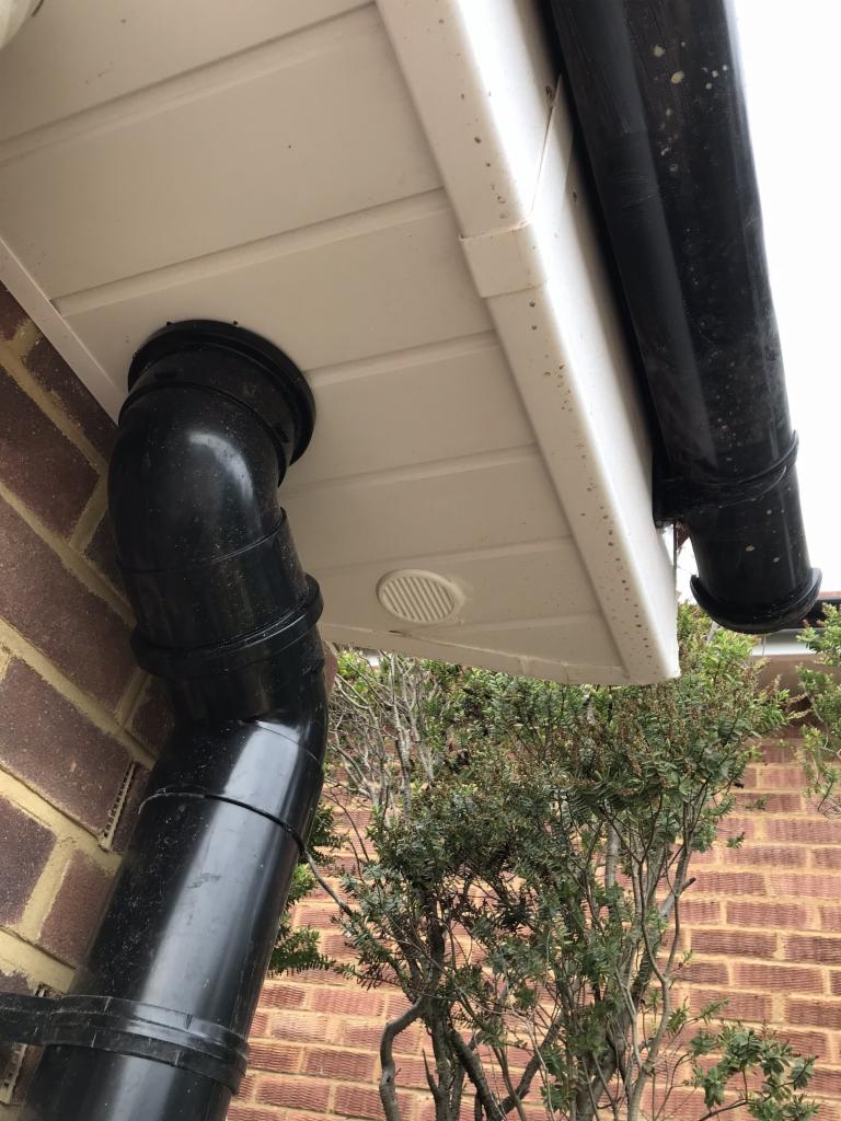 Zamontowana czarna rura spustowa przy okapie z białymi panelami, widoczne zabrudzenia na panelach i rurze, ceglana ściana budynku w tle.
