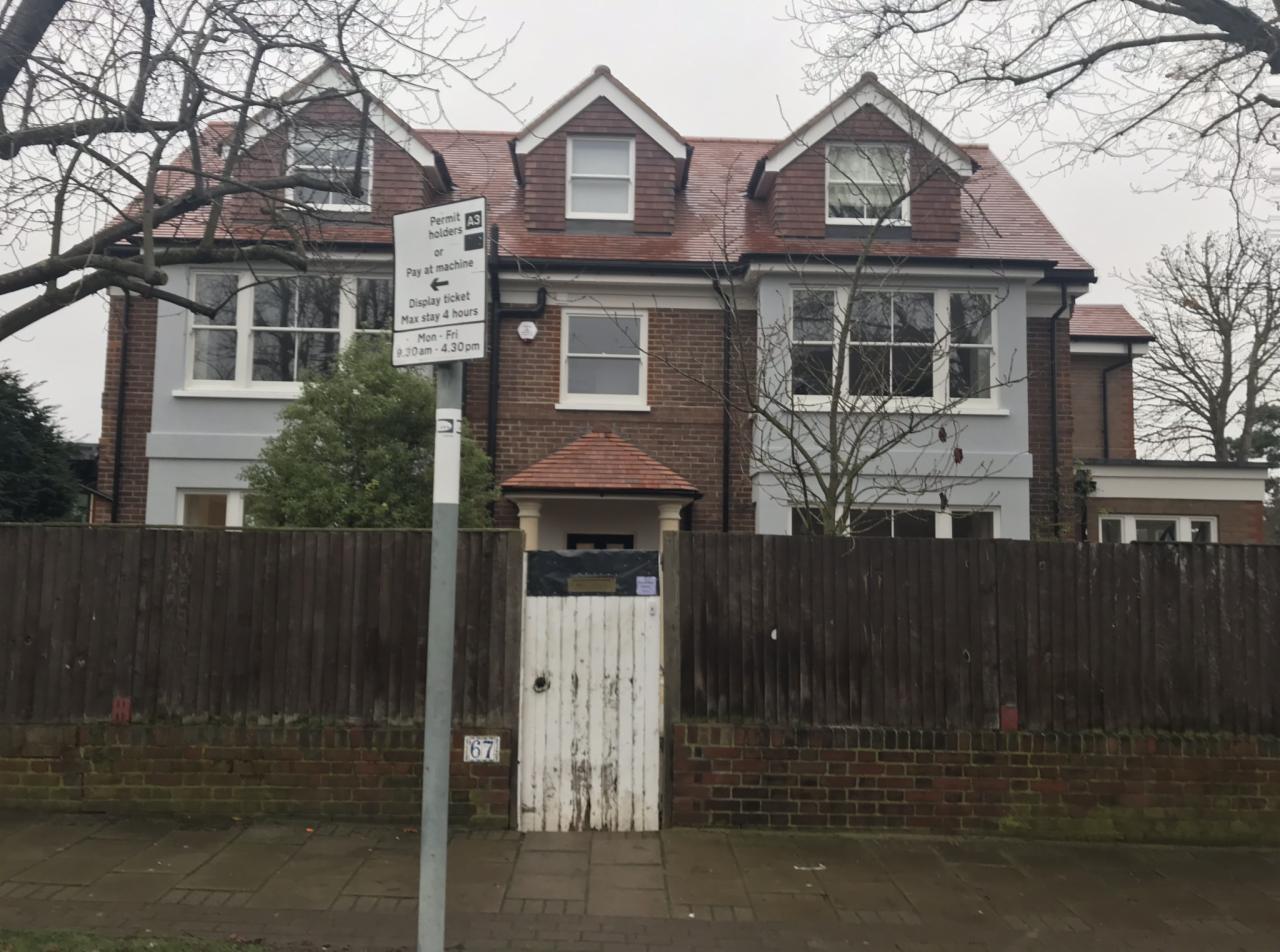 Dwupiętrowy dom z czerwonym dachem i ceglaną fasadą, widoczny zza drewnianego płotu i znaku drogowego z ograniczeniem parkowania.