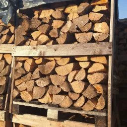 Stos pociętego drewna ułożonego na paletach i zabezpieczonego czarną folią, widoczne słoje drewna i struktura drewna.