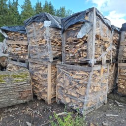 PRYMEK Arkadiusz Kołakowski - Stosy pociętego drewna ułożone w drewnianych skrzyniach na paletach, owinięte folią, z czarną plandeką na górze, na tle drzew i nieba.